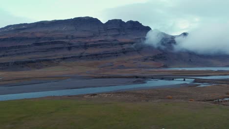 Sheep-grazing-in-a-grass-field-by-a-river-below-a-rock-massif,-Iceland