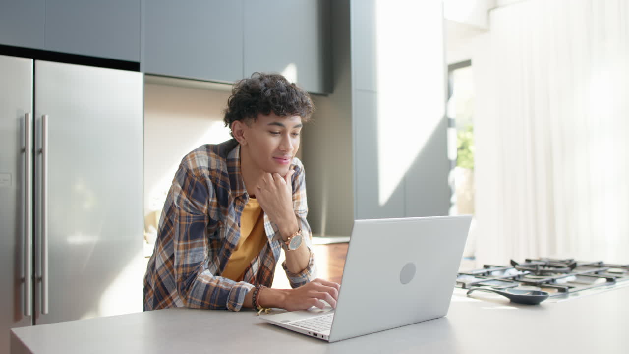 Using laptop and smiling, teenage boy working from home in kitchen