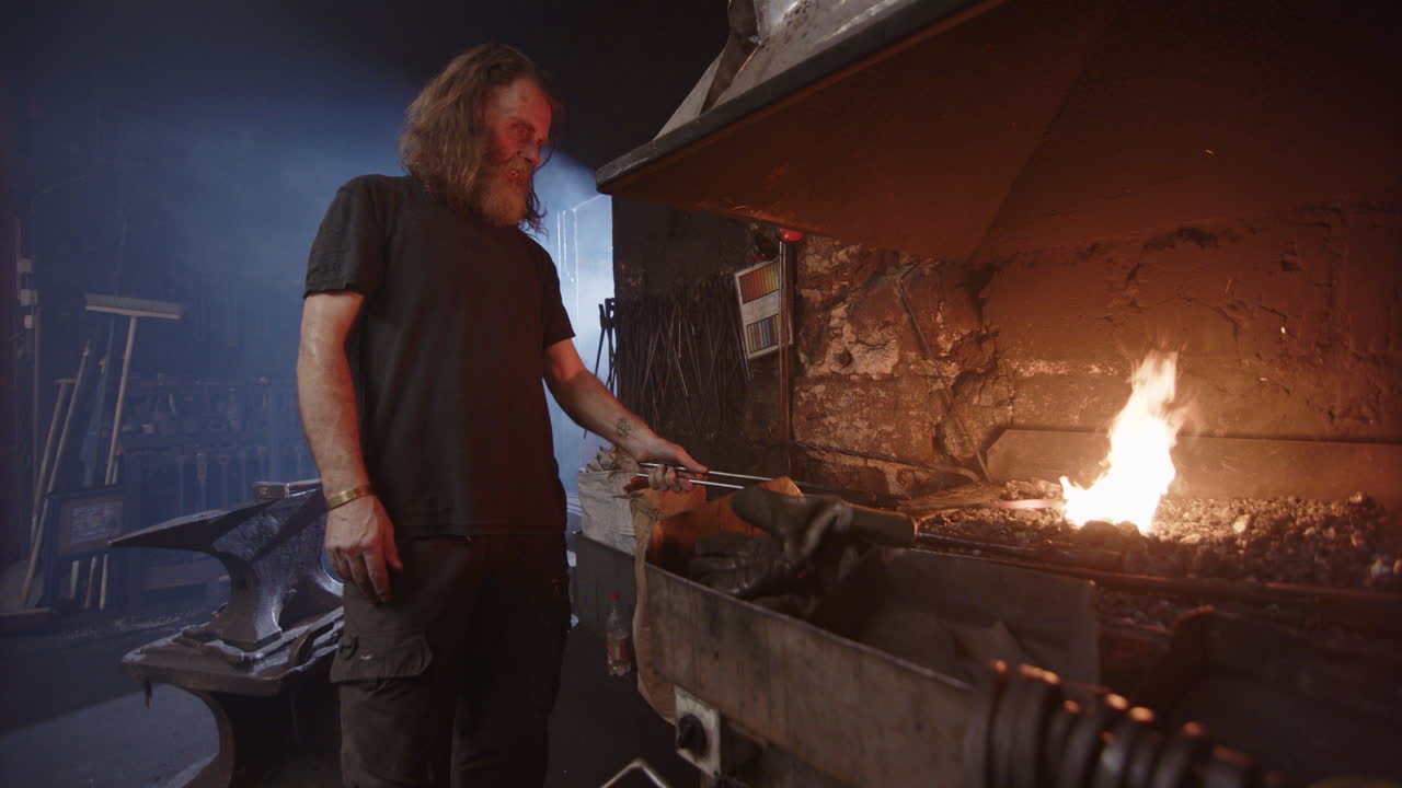 Blacksmith inspects his glowing iron to see if its ready for working on anvil
