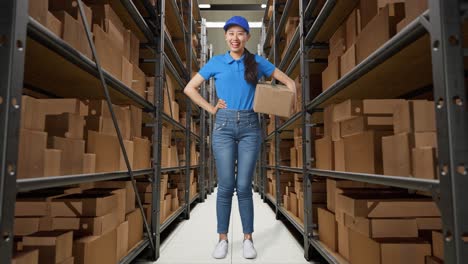 full body of asian female courier in blue uniform putting hand on hip and smiling while delivering a carton in warehouse, pround pose