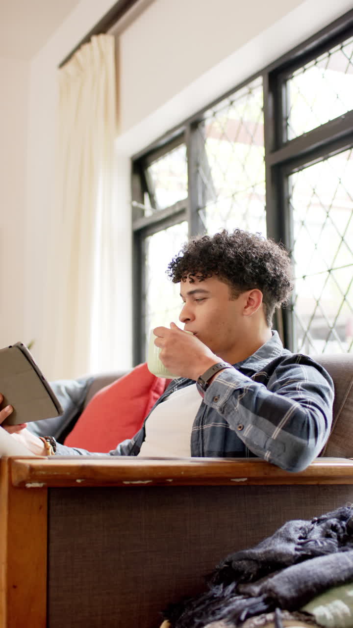 Vertical video of biracial man sitting on couch, using tablet and drinking coffee, slow motion