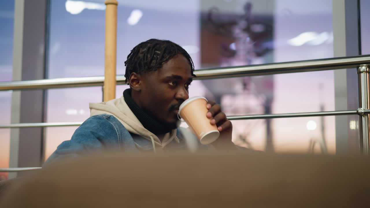 Close up of young man in denim jacket and beige hoodie sitting comfortably in modern cafe during peaceful sunset, looking slightly aside with calm expression, surrounded by soft lighting and warm tones