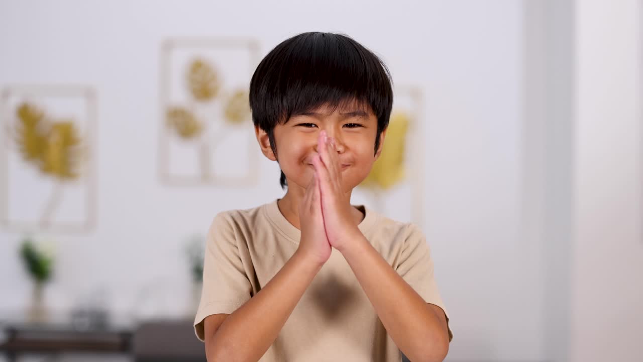 Young Asian boy stands indoors, pressing palms together in a pleading gesture, with varied facial expressions. Soft, even lighting and shallow depth of field highlight subject