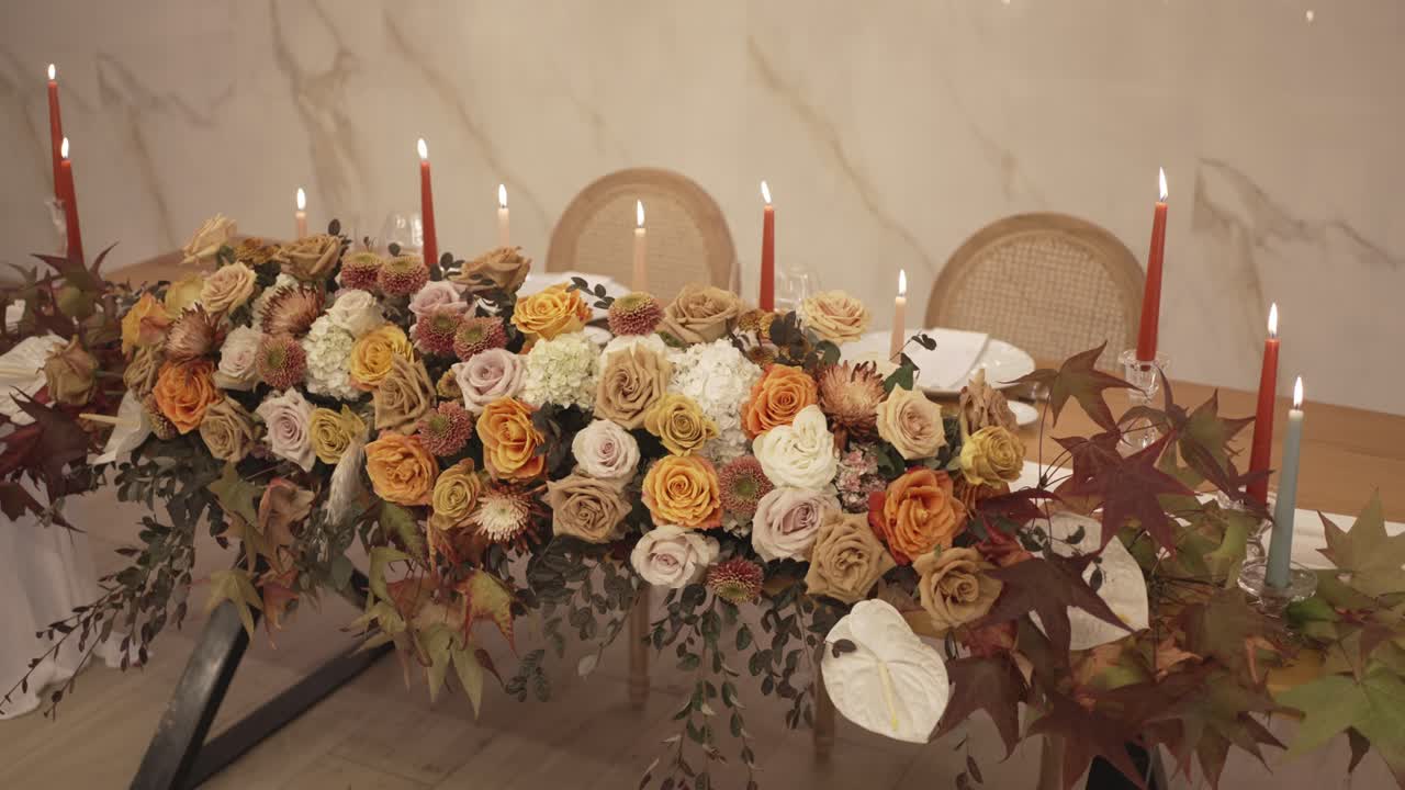 Long table covered in roses foliage and maple leaves with lit red candles and woven chairs behind