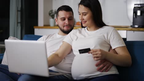 Paar-Erwartet-Ein-Baby,-Sitzt-Zu-Hause-Auf-Dem-Sofa-Und-Kauft-Online-Mit-Dem-Laptop-Ein