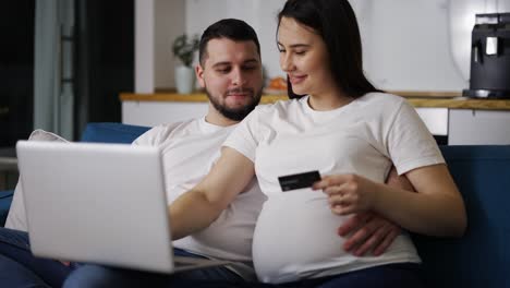 pareja esperando un bebé, sentada en el sofá en casa y comprando en línea usando una computadora portátil