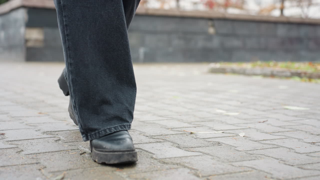 Close up of someone's black trousers and black boots walking slowly toward camera on wet paved urban path, face not visible, capturing calm motion of steps and moody rainy atmosphere