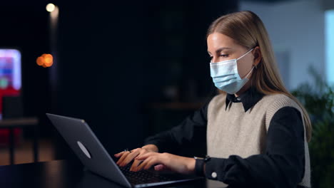 business woman wearing mask opens laptop and working at it. blonde young lady printing something and smiling behind the mask. side view. backdrop in blur.