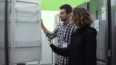 Young-woman-standing-opening-door-of-refrigerator-with-male-consultant-discussing-design-and-quality-before-buying-in-a-consumer-electronics-store.-Discussing-characteristics-with-store-advisor