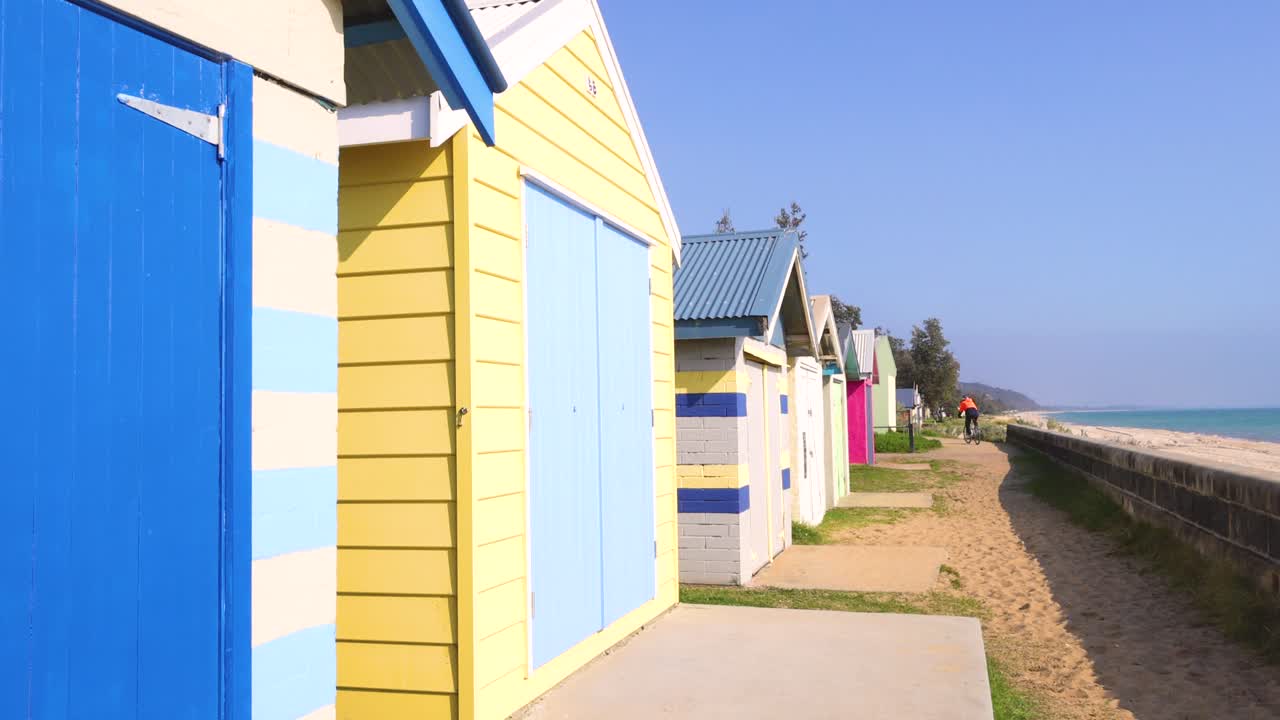Vibrant beach huts line a sandy path under clear skies on Mornington Peninsula, creating a lively and picturesque coastal scene