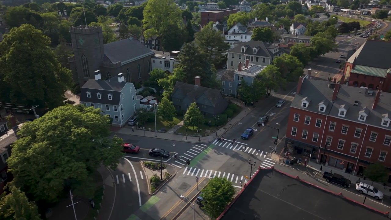 Witch House gambrel roof rises among dense green trees near crosswalk-marked intersection, cars crossing past brick storefronts along historic Salem, Massachusetts street. Aerial drone view