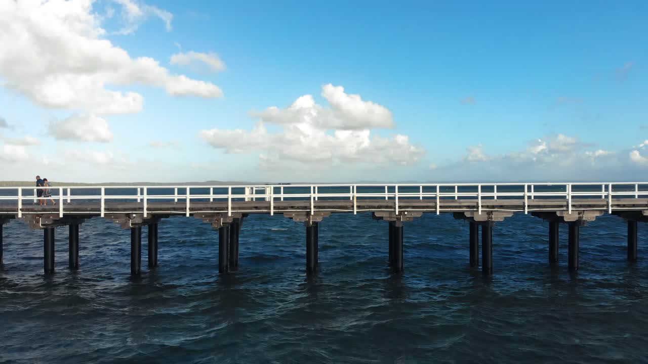 아름다운 열대의 푸른 하늘 아래 깊고 짙은 푸른 물이 있는 광대한 바다를 따라 뻗어 있는 길고 오래된 나무 부두의 왼쪽에서 오른쪽으로 빠르게 미끄러지는 장면