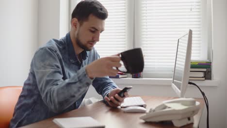 Young-office-worker-using-his-phone-at-the-office-sitting-at-the-table-with-computer,-phone-and-drinking-coffee-or-tea-from-his