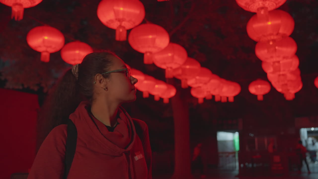 Farolillos rojos colgando sobre una mujer contemplando la noche, viajera urbana bajo un dosel de farolillos rojos brillantes cerca de la entrada de un templo, luz cálida reflejada en sus gafas y bufanda, ambiente de festival tranquilo con sutileza.