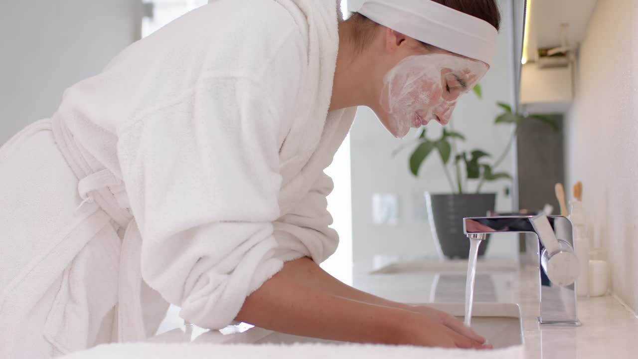 Washing face with cleanser, woman in bathrobe enjoying skincare routine at home