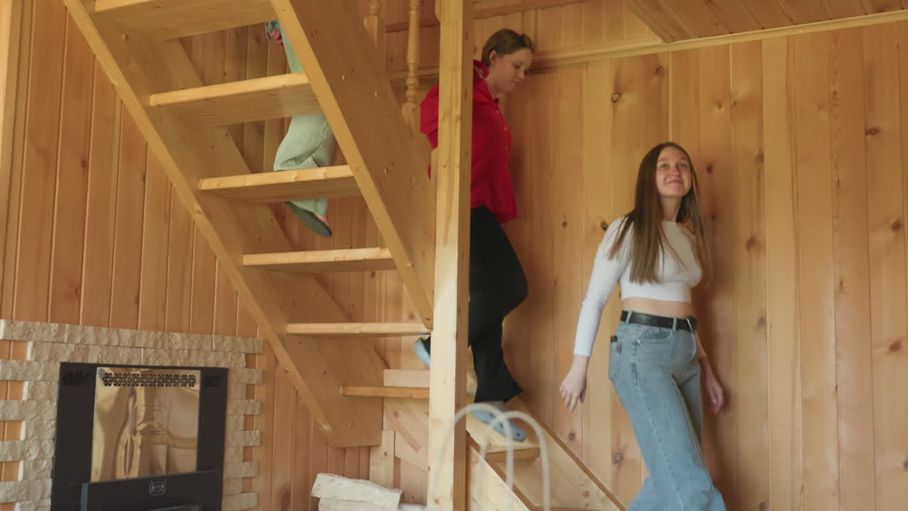 two women step down wooden staircase inside rustic cabin, soft lighting highlights wooden walls and structure, casual attire and relaxed pace suggest calm atmosphere