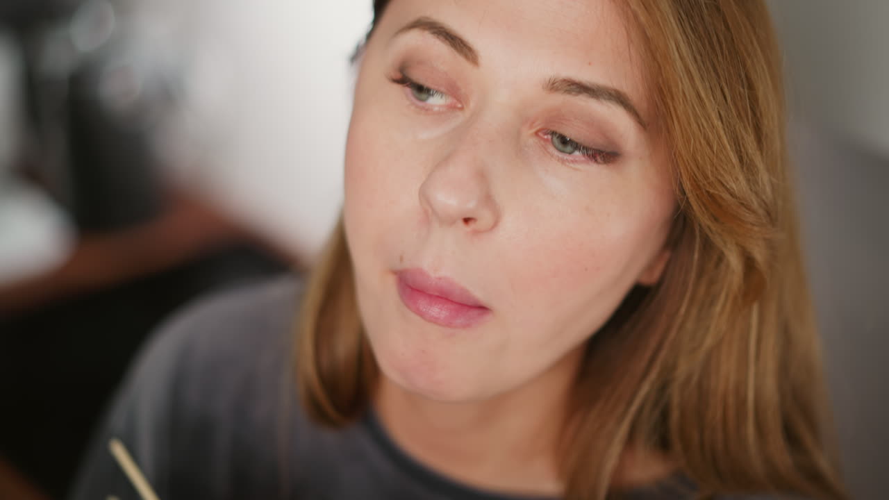 Lady eating food using chopsticks while distracted, looking away thoughtfully in bright modern kitchen, blurred appliances and kitchen items softly visible in background