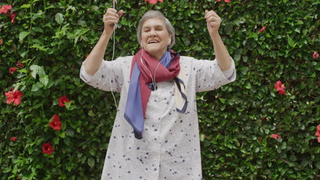 retrato de una mujer caucásica mayor bailando brazos juguetones levantados disfrutando escuchando música en auriculares al aire libre fondo del jardín mujer vieja animada