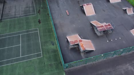 Vista-Aérea-Volando-Sobre-La-Rampa-Del-Parque-De-Patinaje-Cercado-Y-La-Cancha-De-Tenis-En-Un-Patio-De-Juegos-Vacío-Y-Cerrado