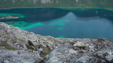 female wanderlust sitting on rocky hill salberget with pet dog overlooking calm blue lake in norway