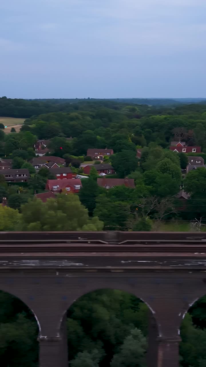 Vista aérea vertical con dron de un viaducto ferroviario en Hertfordshire al atardecer. El dron se mueve hacia la izquierda y capta las vías, los arcos y el cielo azul del atardecer sobre los árboles circundantes y el campo abierto.