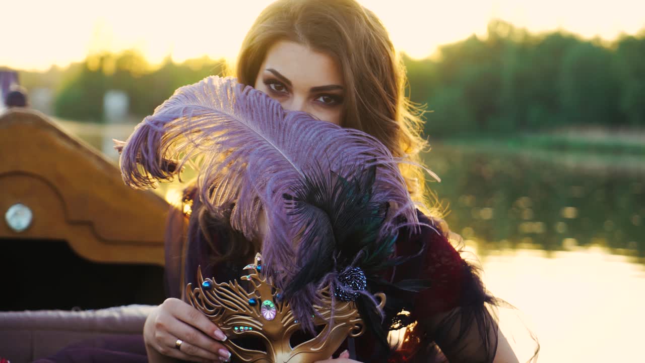 Beautiful woman posing into a camera while floating in a boat and closing her face with feather at sunset. Pretty female holding carnival mask with purple feather. Close-up