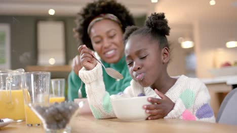 happy african american mother and daughter eating cereal with milk in kitchen, slow motion