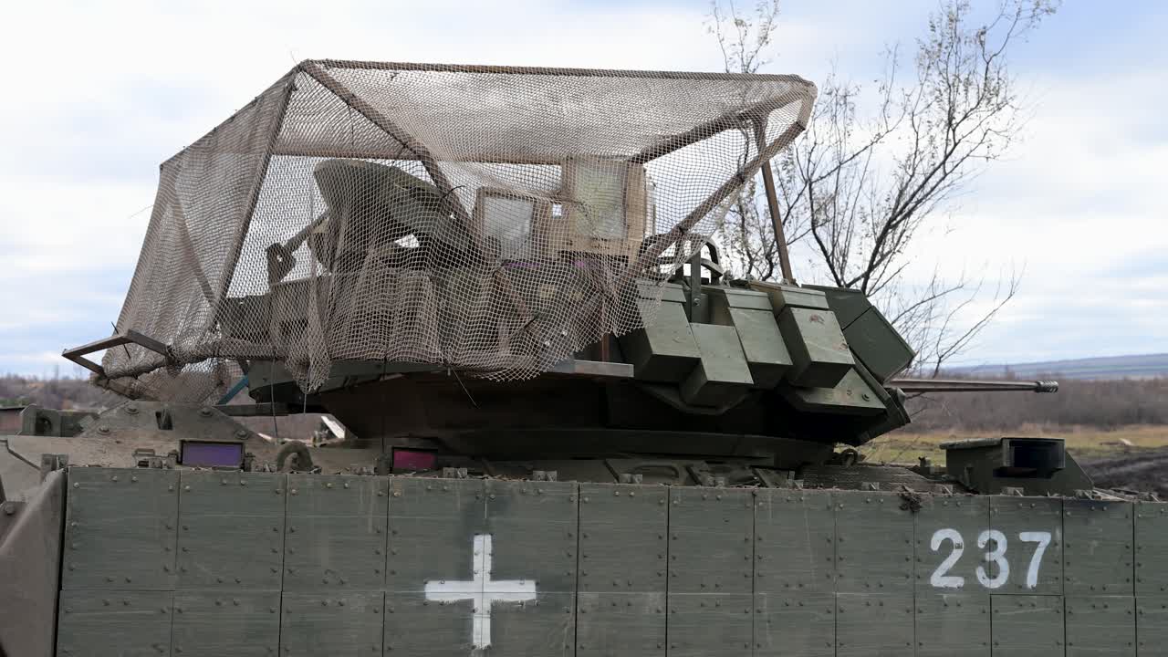 Mentre la sua torretta ruota, un veicolo da combattimento della fanteria M2 Bradley logorato dalla battaglia giace ricoperto di fango sul fronte ucraino, contrassegnato per servizio medico e corazzato contro la costante minaccia dei droni