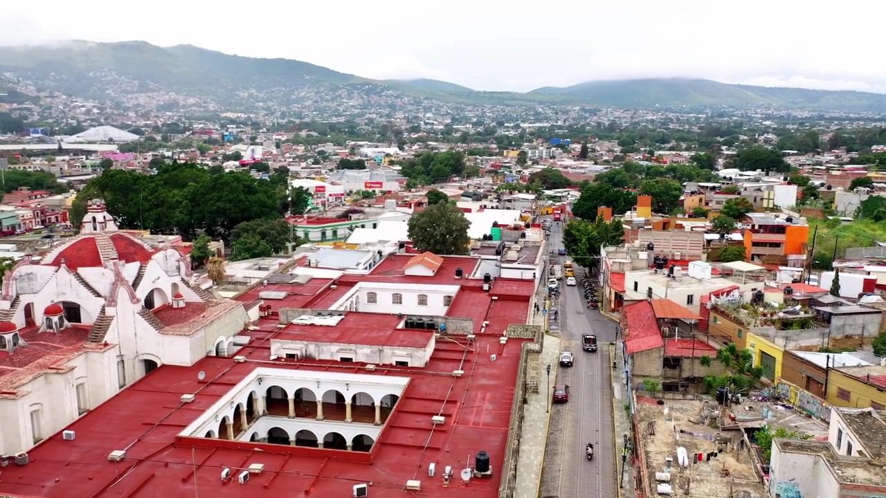 Ripresa aerea con drone di un colorato quartiere della città di Oaxaca con edifici densi, architettura vivace, collina verde e paesaggio urbano durante il giorno nuvoloso.