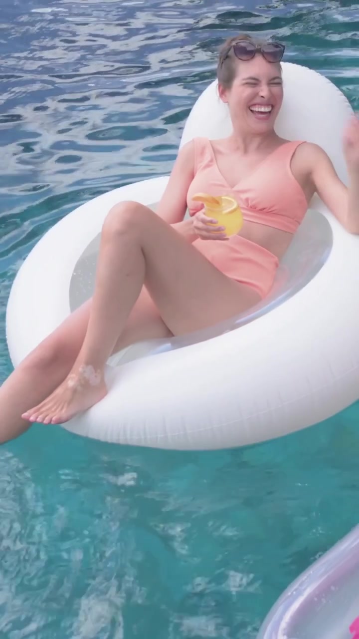 Vertical video: Laughing woman in peach swimsuit lounging on float, reaching for offered yellow cup