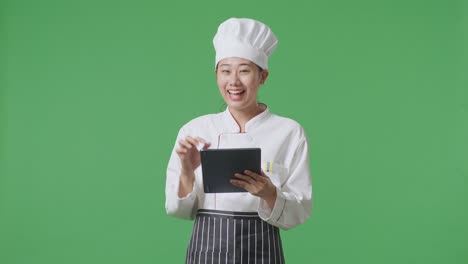 Asian Woman Chef Using A Tablet And Pointing To Side While Standing In The Green Screen Background Studio