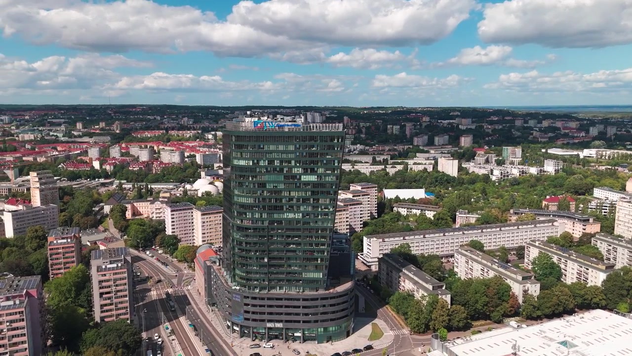 Imagem aérea de drone de um arranha-céu contemporâneo cercado por blocos residenciais e espaços verdes sob um céu azul com nuvens brancas.