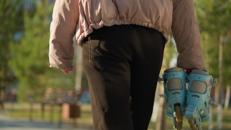 close-up of a person holding a pair of rollerblades, wearing black trousers and a peach jacket, walking through a park with a blurred background of trees