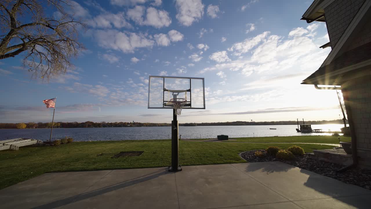 canestro da basket su un campo accanto a una casa di lusso sul lago con la bandiera americana che sventola sullo sfondo, perdite di luce solare