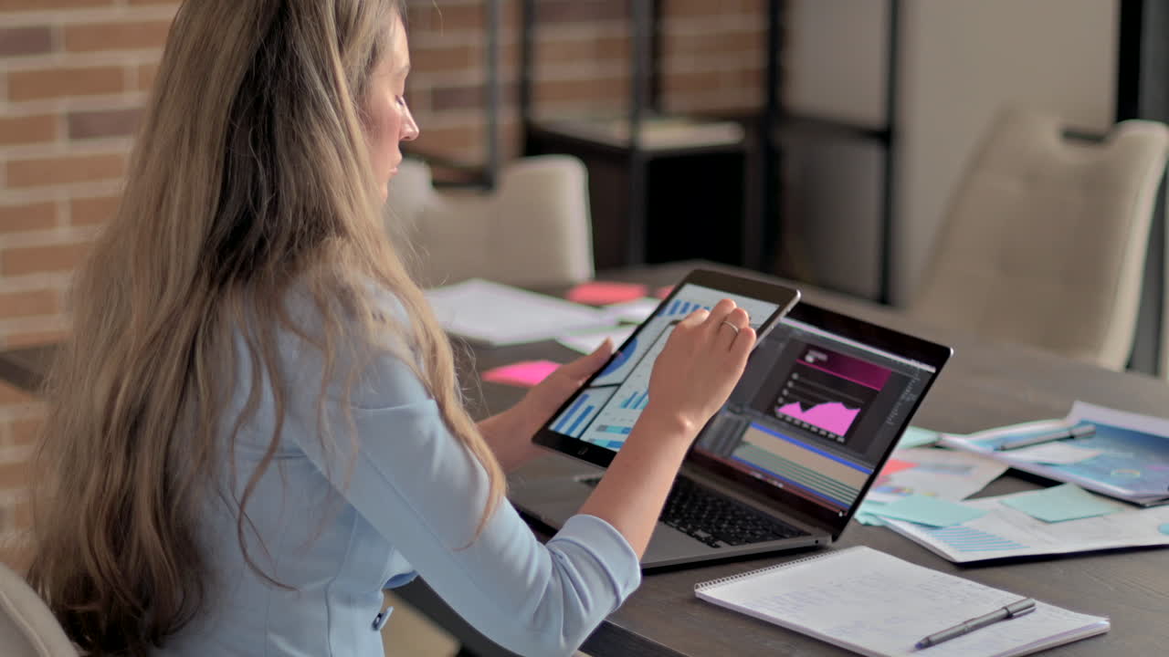 Donna con blazer blu che lavora su un tablet davanti a un laptop, a una scrivania, in un ufficio