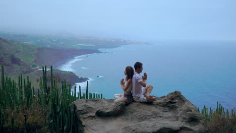 a man and woman atop a mountain sit on a rock back to back, engaging in meditation and yoga while absorbing the ocean's serenity