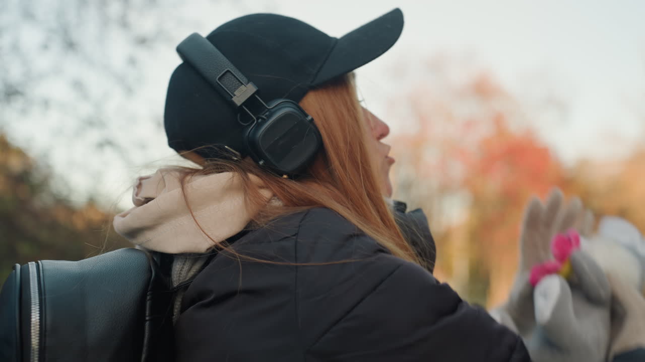 Donna con zaino e cuffie in un parco autunnale che reagisce a un sospetto tentativo di scippo, che si protende verso la custodia della chitarra, con berretto, sciarpa e cappotto, profilo posteriore e laterale spontaneo, alberi e foglie autunnali con effetto bokeh morbido