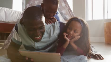 video of happy african american father, son and daughter lying in tent and using tablet together