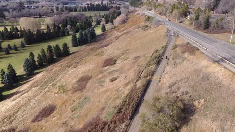 3-4-Antenne-Fliegen-Vorwärts-über-Den-City-Valley-River-Golfplatz-Neben-Einem-Ungepflegten-Park,-Während-Die-Menschen-Beim-Joggen-Auf-Dem-Betonierten-Weg-Am-Straßenrand-Soziale-Distanz-Voneinander-Halten