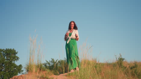Young woman walking down grassy hillside carrying bubble solution, green dress flowing in gentle breeze under clear blue sky, warm sunset light casting long shadows over tranquil river valley