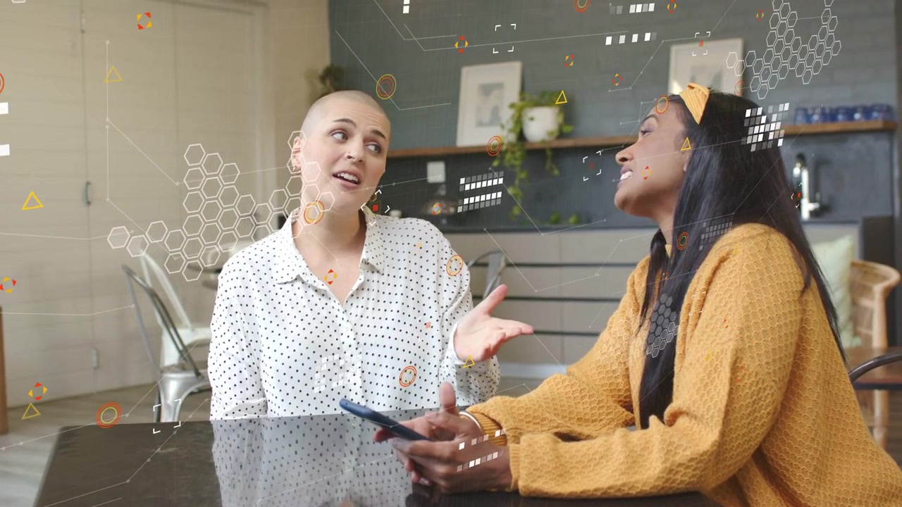 Woman in mustard holding phone and speaking to dotted blouse, gesturing while sharing tech overlays