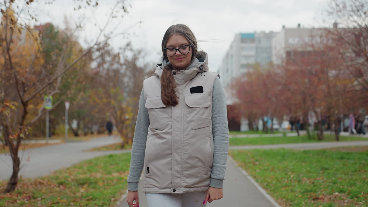 Elegant lady wearing glasses and ash jacket walking with a smile in urban park setting with autumn trees, green grass, moving cars, and residential buildings in background