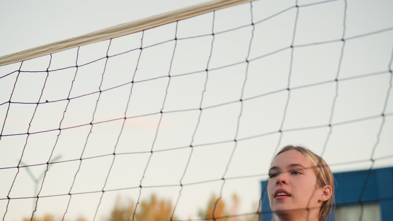 젊은 여성 운동선수가 점프하는 클로즈업, 집중과 결단력으로 배구 타격 준비, 승리를 보여주는, 배경에 배구 망과 멀리서 볼 수 있는 건물