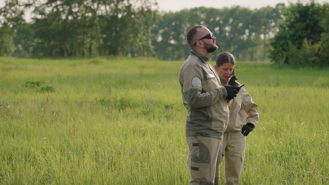 wife on call stands close to husband scanning sky horizon while he tracks aerial target with device tools in grassy field under clear sky at sunset research operation teamwork