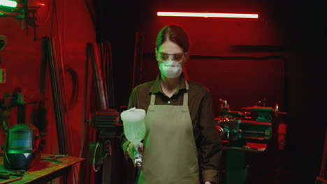 caucasian woman in goggles, respiratory mask and apron working with pulverizator in workshop