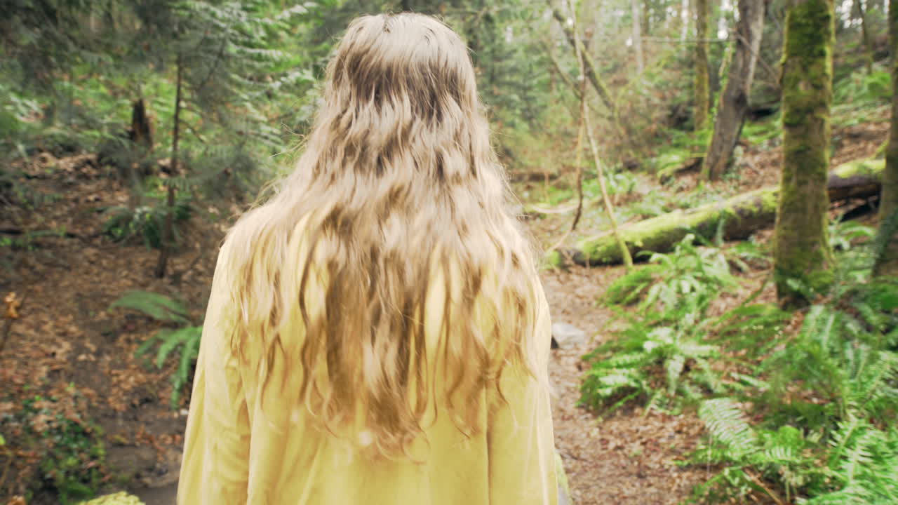 A young woman wearing a bright yellow jacket walks over a bridge and through a green mossy forest, tracking shot from behind
