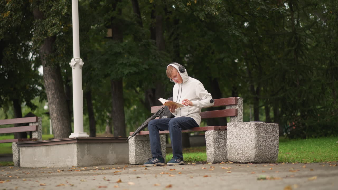 구름이 낀 나무 아래 벤치에 앉아 태블릿으로 음악을 듣고 있는 노인, 근처에 가로등, 젖은 보도와 흩날리는 낙엽, 옆에는 캐주얼한 청바지와 백팩, 은은한 흐린 조명, 재생 목록 스크롤 중