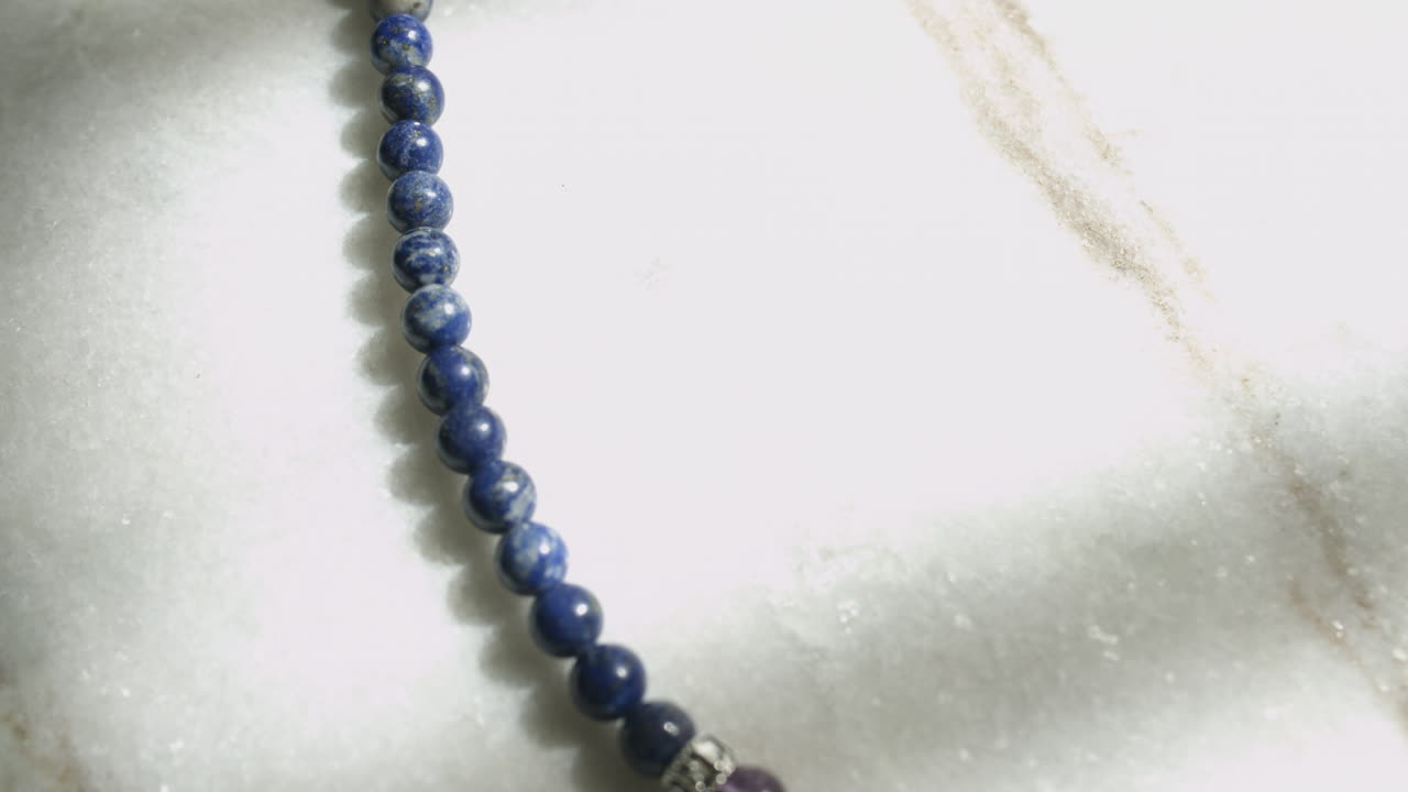 Macro carrellata di perle di lapislazzuli blu da una mala di preghiera tibetana che poggiano su una lastra di marmo bianco, ripresa in UHD