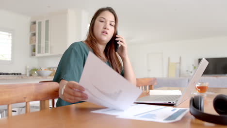 mujer biracial ocupada con un documento, usando una computadora portátil y hablando por teléfono en casa, en cámara lenta