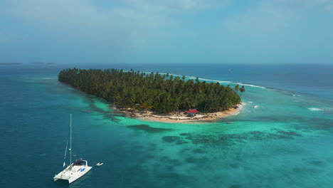 Sailboat-Floating-Near-Remote-Tropical-Island-With-Green-Palm-Trees,-Surrounded-By-Bright-Blue-Sea